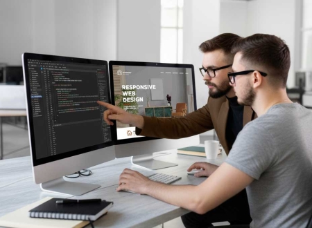 Zwei Männer besprechen eine Website am Schreibtisch mit zwei Bildschirmen, einer zeigt HTML-Code, der andere das fertige Design